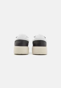 Sneaker in pelle bianca con accenti neri, con una suola testurizzata e una forma arrotondata. Branding visibile sul tallone. Design minimalista.