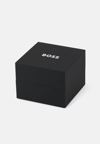 Boîte carrée noire avec une texture lisse et un couvercle, portant le nom de la marque "BOSS" imprimé en blanc sur le dessus.
