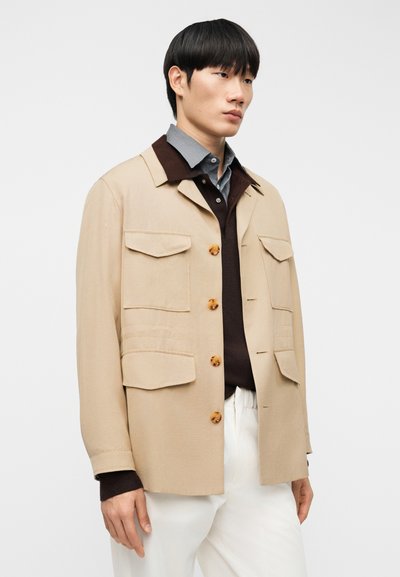 Μπεζ σακάκι με δύο τσέπες στο στήθος, καφέ γιακά, μεγάλα κουμπιά και άνετη εφαρμογή, φορεμένο πάνω από σκούρο πουλόβερ και ανοιχτόχρωμο παντελόνι.