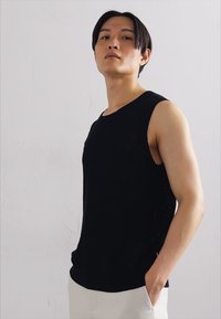 Black sleeveless top made of textured fabric, featuring a round neckline and slightly relaxed fit. Model stands against a neutral background.