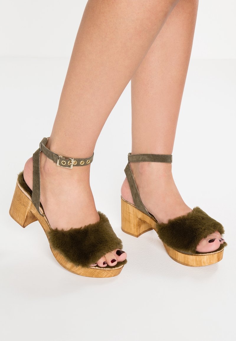 Pieds portant des sandales à talon bloc ouvert avec des brides en fourrure vert olive et des fermetures à boucle à la cheville sur un fond clair.