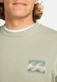 Lichtgroene sweatshirt met een ribbelnek en een vierkante graphic met golfpatronen in blauw en wit. Zacht, gestructureerd stof.
