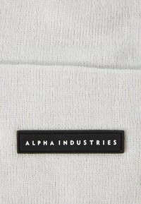 Világosszürke kötött sapka fekete téglalap alakú folttal, amelyen fehér szöveggel olvasható az "ALPHA INDUSTRIES". Az anyag puhanak tűnik, bordázott textúrával.