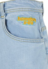 Gaisni zili džinsi ar izšūtu "homeboy x-tra" logotipu dzeltenā un zilā krāsā, kas aprīkoti ar priekšējo kabatu un redzamām šuvju detaļām.