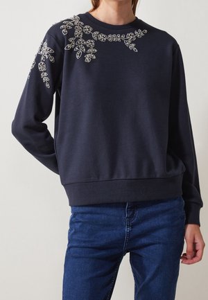 Femme portant un pull bleu marine foncé avec des détails brodés de perles florales sur l'épaule et le haut de la poitrine, associé à un jean bleu.