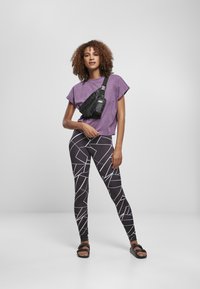 T-shirt cropped viola, leggings neri con motivo geometrico bianco, borsa a tracolla nera e sandali neri con suola piatta.