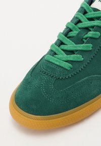Zaļie zamšas sporta apavi ar teksturētu virsu, slāņotu šuvju dizainu un atbilstošām auklām, ar gumijas zoli.