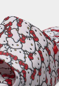 Λευκό αντικείμενο καλυμμένο με επαναλαμβανόμενα πρόσωπα Hello Kitty με κόκκινα φιόγκους και κίτρινες μύτες, με μια μικρή κόκκινη καρφίτσα στη μία πλευρά.