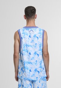 Mann iført blått og hvitt batikkfarget ermeløst drakt og matchende shorts, stående med ryggen mot på en ensfarget grå bakgrunn.