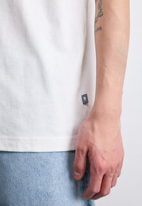 Tricou alb din bumbac, cu mâneci scurte, având o etichetă mică și închisă la culoare pe cusătura laterală; purtat cu blugi din denim bleu deschis.