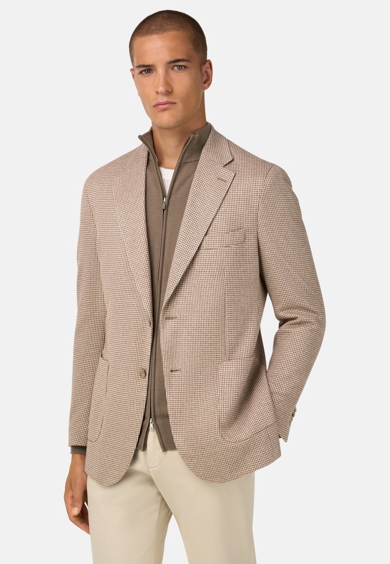 Blazer beige a fantasia con rever a punte, due tasche frontali e bottoni dorati, indossato sopra una camicia marrone con zip. Vestibilità tailored.