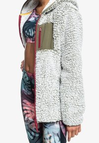 Fleecejakke med en textured hvid krop, olivengrøn brystlomme og lyserød lynlås. Bæres med mønstrede leggings med tropiske designs.