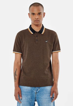 Polo shirt marron à motifs avec un col noir et des accents dorés et un petit logo sur la poitrine. Porté avec un jean bleu clair.