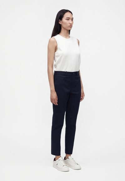 Femme debout face à l'objectif, portant un haut blanc sans manches, un pantalon slim foncé et des baskets blanches sur un fond blanc uni.