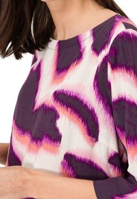 Lila- und rosa gemusterte Bluse mit lockerer Passform, Rundhalsausschnitt und Dreiviertelärmeln aus einem glatten Stoff mit leicht glänzender Textur.