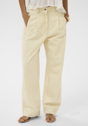 Vrouw met losse beige broek met voorvouwen, witte blouse, beige sandalen en gouden armband, handen in de zakken.