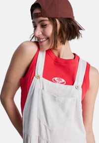 Salopette en denim blanc avec grande poche avant et boutons en bois, portée sur un débardeur rouge avec un logo blanc.