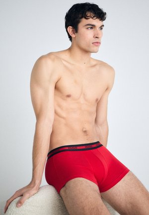 Piros boxer alsónemű fekete márkázott derékkal, testhezálló kialakítással, sima textúrával és egyszínű dizájnnal. A modell ülve, meztelen törzzsel.
