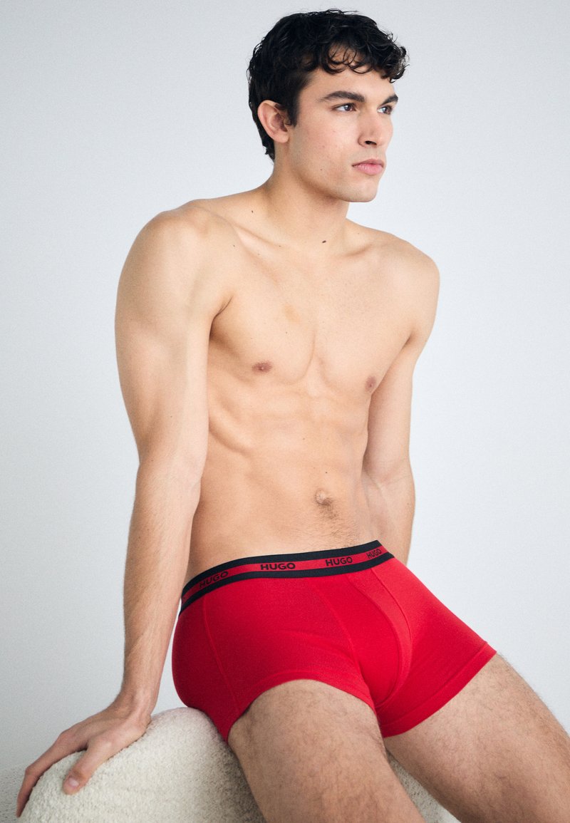 Røde boxershorts med en sort mærkebånd i taljen, med en tætsiddende design, glat tekstur og ensfarvet. Modell sitter med bar overkropp.
