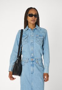 Világoskék farmering gombos zárással és mellzsebekkel, világoskék farmer szoknyával párosítva, fekete, redőzött kézitáskával. Fekete napszemüveg viselve.