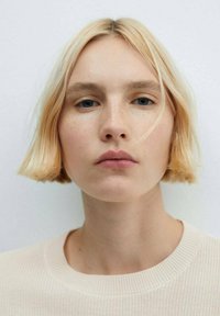 Capelli biondi, bob alla lunghezza del mento con onde morbide; pelle chiara con lentiggini; maglione neutro con scollo a costine; sfondo tenue.