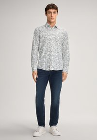 Chemise à manches longues de couleur claire avec un motif géométrique, assortie à un jean en denim foncé et des baskets blanches, présentant une coupe décontractée.