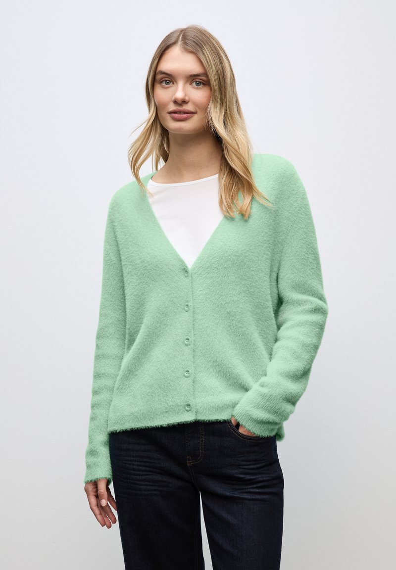 Jeune femme portant un cardigan vert menthe en laine pelucheuse sur un chemisier blanc et un jean foncé, debout devant un fond gris clair uni.