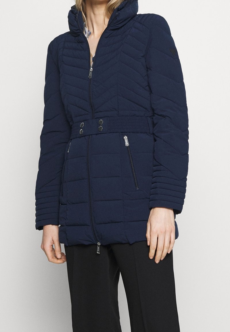 Veste doudoune bleu marine avec fermeture éclair à l'avant, capuche, motif matelassé en chevrons, ceinture ajustable à la taille et poches latérales zippées. Tissu lisse et isolant.