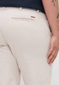 Beige pants with a smooth, textured fabric. Features a back pocket with a brown leather accent, visible stitching, and a fitted design.