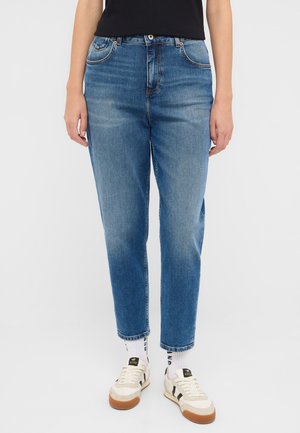 Jasnoniebieskie dżinsy o prostym kroju, z pięcioma kieszeniami, zapięciem na guzik i zamek błyskawiczny oraz lekkim przetarciem. Noszone z beżowymi trampkami.