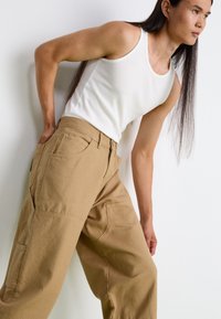 Tan wide-leg pants made of durable material, featuring side pockets, a zip and button closure, paired with a white fitted tank top.