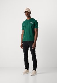 Grønn bomull T-skjorte med logo, korte ermer, sammen med svarte jeans og beige sneakers. Uformell bekledning. Enkelt design.