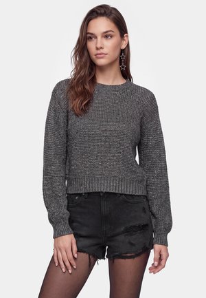 Ung kvinde iført en mørkegrå strikket sweater, sorte revnede denimshorts, gennemsigtige sorte strømpebukser og stjerneformede hængeøreringe.
