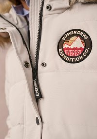 Weiße Pufferjacke mit einem Reißverschluss vorne, versehen mit einem runden Aufnäher mit einem Berg und dem Text "Superdry Everest Expedition Co".