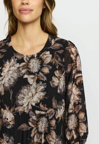 Kvinne iført en svart bluse med store beige og brune blomstertrykk og rund hals mot en ensfarget lys bakgrunn.