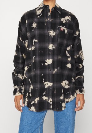 Camisa de cuadros negra y gris con botones y salpicaduras blancas de lejía, llevada con vaqueros azules, mostrada desde los hombros hasta las caderas.