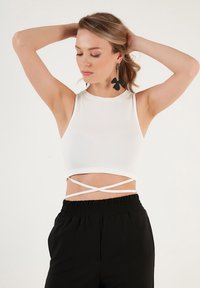 Bijela rebrasta crop majica s bezrukavim dizajnom i detaljem u obliku križa na struku, u kombinaciji s crnim hlačama s elasticom u struku.