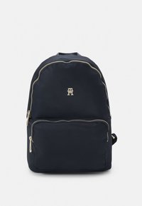 Tamsiai mėlynas kuprinės su lygiu paviršiumi, aukso spalvos užtrauktuku, priekiu kišenėle ir logotipu viršutinėje dalyje.