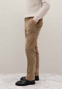 Braune Cordhosen mit gerader Passform, ausgestattet mit Seitentaschen und strukturiertem Stoff. Kombiniert mit schwarzen Schuhen, präsentiert auf einer hellen Oberfläche.