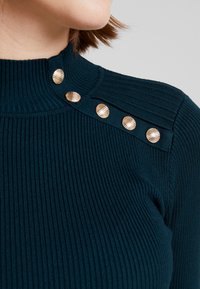 Geribde teal-col top met col en goudkleurige decoratieve knopen op de schouder, die textuur en details aan de stof toevoegen.