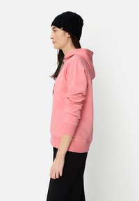 Pink hoodie with a kangaroo pocket and drawstring hood, made of soft fabric. Worn with a black beanie, side view against a plain background.