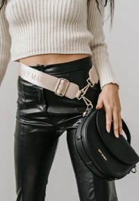 Sac à bandoulière en cuir noir au forme arrondie, orné de quincaillerie métallique et de texte embossé. Associé à un pantalon noir moulant et brillant et un pull en crème à côtes.