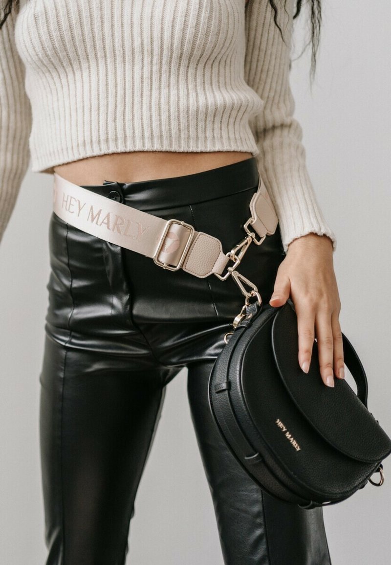 Sac à bandoulière en cuir noir au forme arrondie, orné de quincaillerie métallique et de texte embossé. Associé à un pantalon noir moulant et brillant et un pull en crème à côtes.