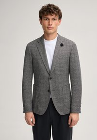 Grijze geruite blazer met een getailleerde pasvorm, twee knopen, notch revers en een enkele bloemenaccent op de revers. Draagt over een wit t-shirt.
