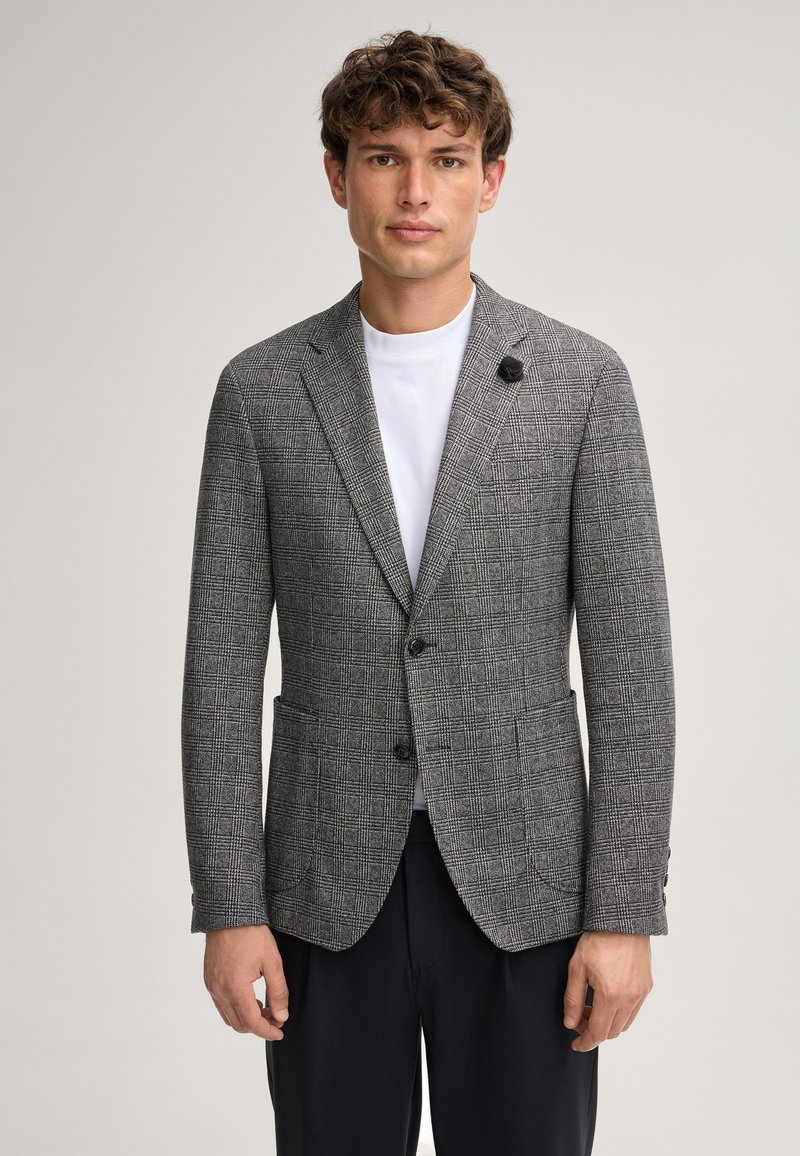 Blazer grigio a quadri con vestibilità su misura, due bottoni, revers a punta e un singolo fiore decorativo sul bavero. Indossato sopra una t-shirt bianca.
