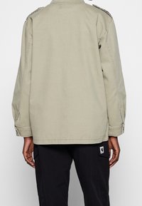 Light olive green cotton jacket with a relaxed fit, featuring shoulder epaulettes and buttoned cuffs. Includes a logo patch on the lower pocket.