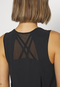 Haut sans manches noir avec un dos en maille transparente, des bretelles diagonales et un ourlet arrondi. Tissu doux avec une coupe ample.