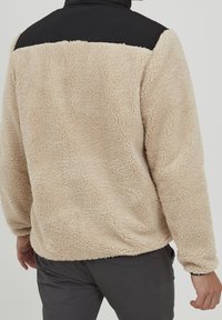Beige fleece trui met een zwarte nylon schouderpartij. Heeft een textuur, zachte stof en lange mouwen met elastische manchetten.