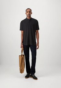 Black polo shirt, short sleeves, flat-knit collar, dark jeans, holding a brown tote bag, standing on a neutral background.