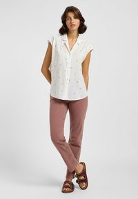 Camisa blanca de manga corta con cierre de botones, que presenta un sutil patrón a rayas y pequeños acentos florales; combinada con pantalones rosas y sandalias.
